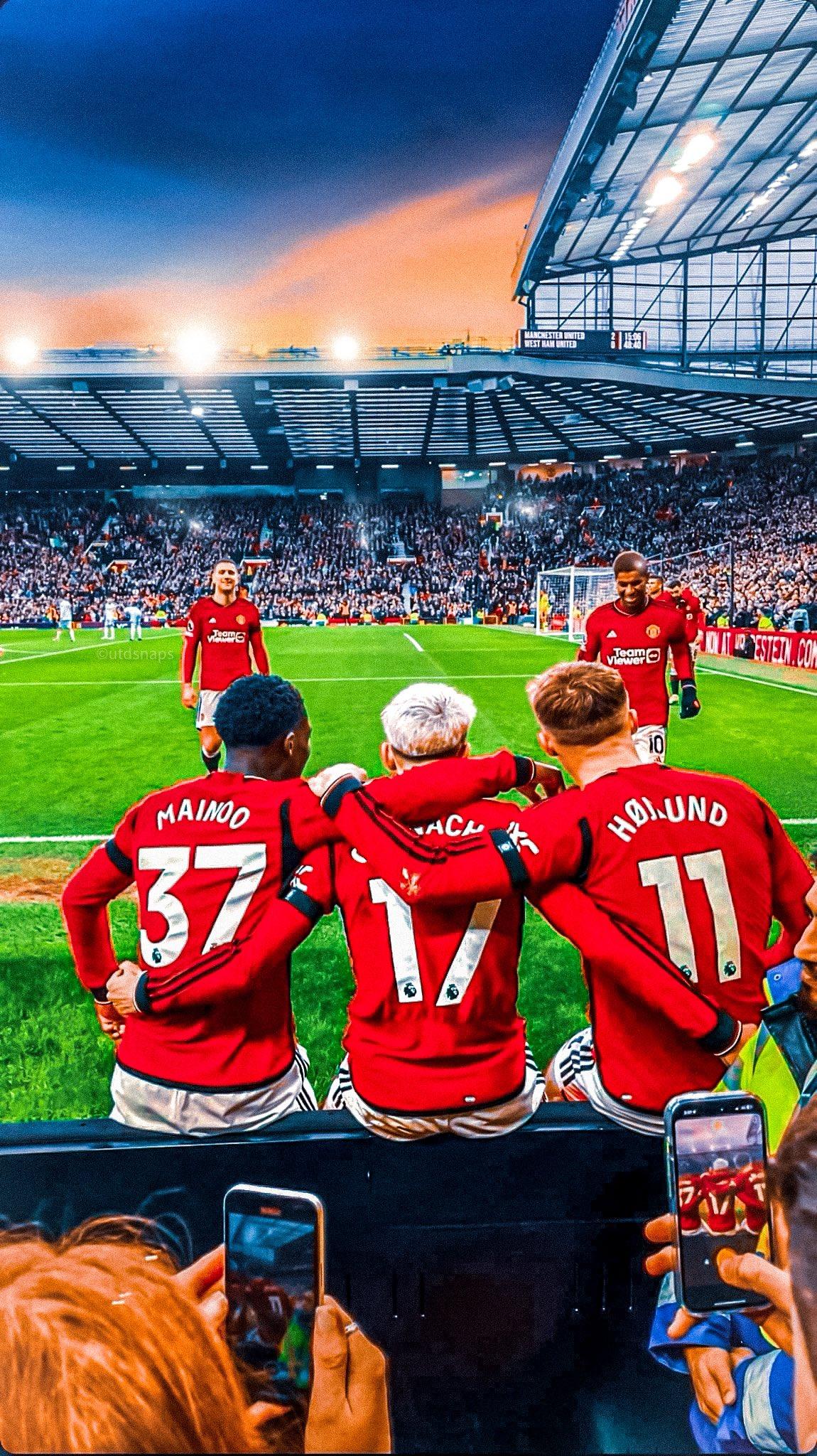 Manchester United players celebrate a goal at Old Trafford as fans capture the moment on their phones. The scoreboard in the background shows Manchester United leading against West Ham United. The sunset creates a vibrant backdrop, enhancing the lively atmosphere of the stadium.