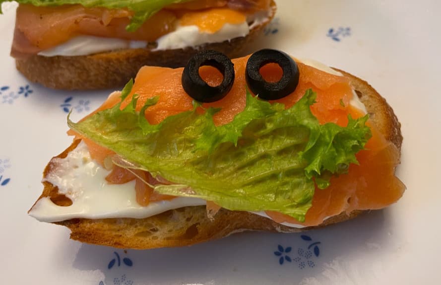 A piece of sourdough with smoked salmon on it. On top of the salmon, a piece of letter shaped like a smile with olives lying above the lettuce to represent eyes.