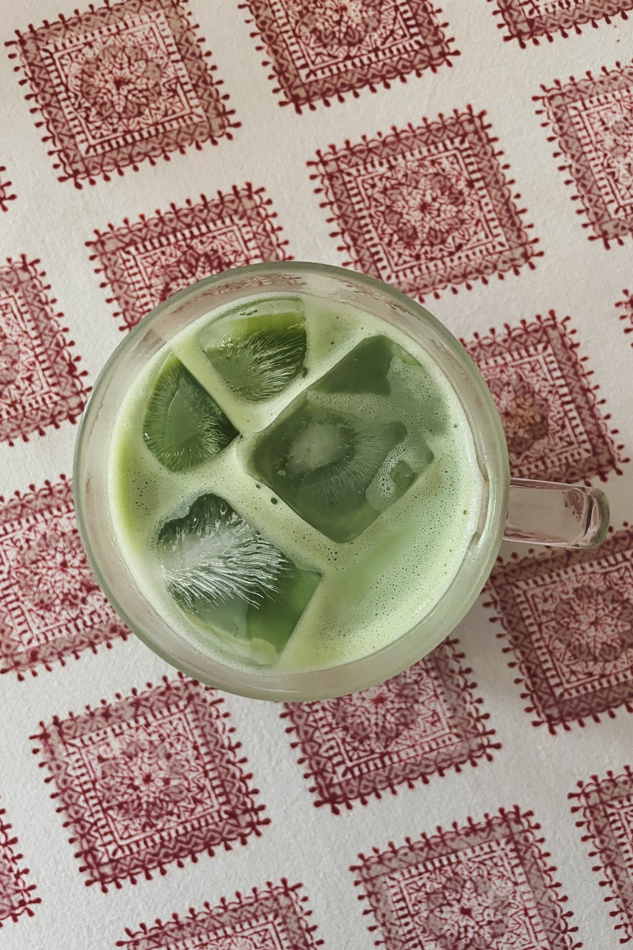 A top-down view of a glass of iced matcha latte placed on a white tablecloth with a red, intricate square pattern. The latte has a light green color and is filled with large ice cubes.