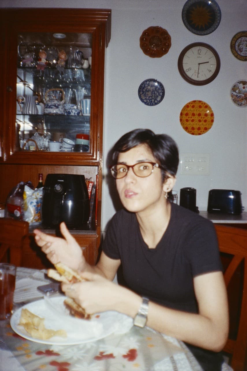 Woman wearing glasses and black shirt eating at table with wooden cabinet and decorative clocks on wall in vintage film photograph