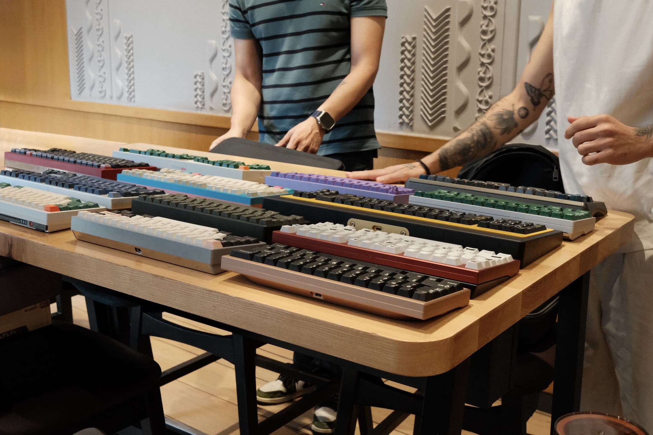 Group of people examining multiple mechanical keyboards with colorful keycaps displayed on wooden table at meetup event