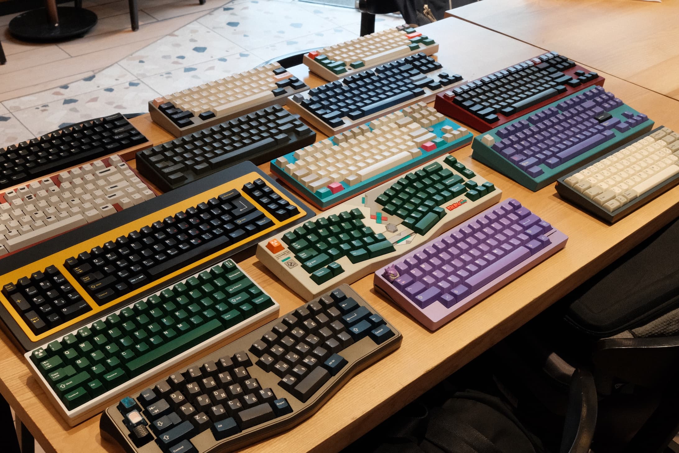 Collection of mechanical keyboards with various colorful keycaps and cases displayed on wooden table at keyboard meetup event